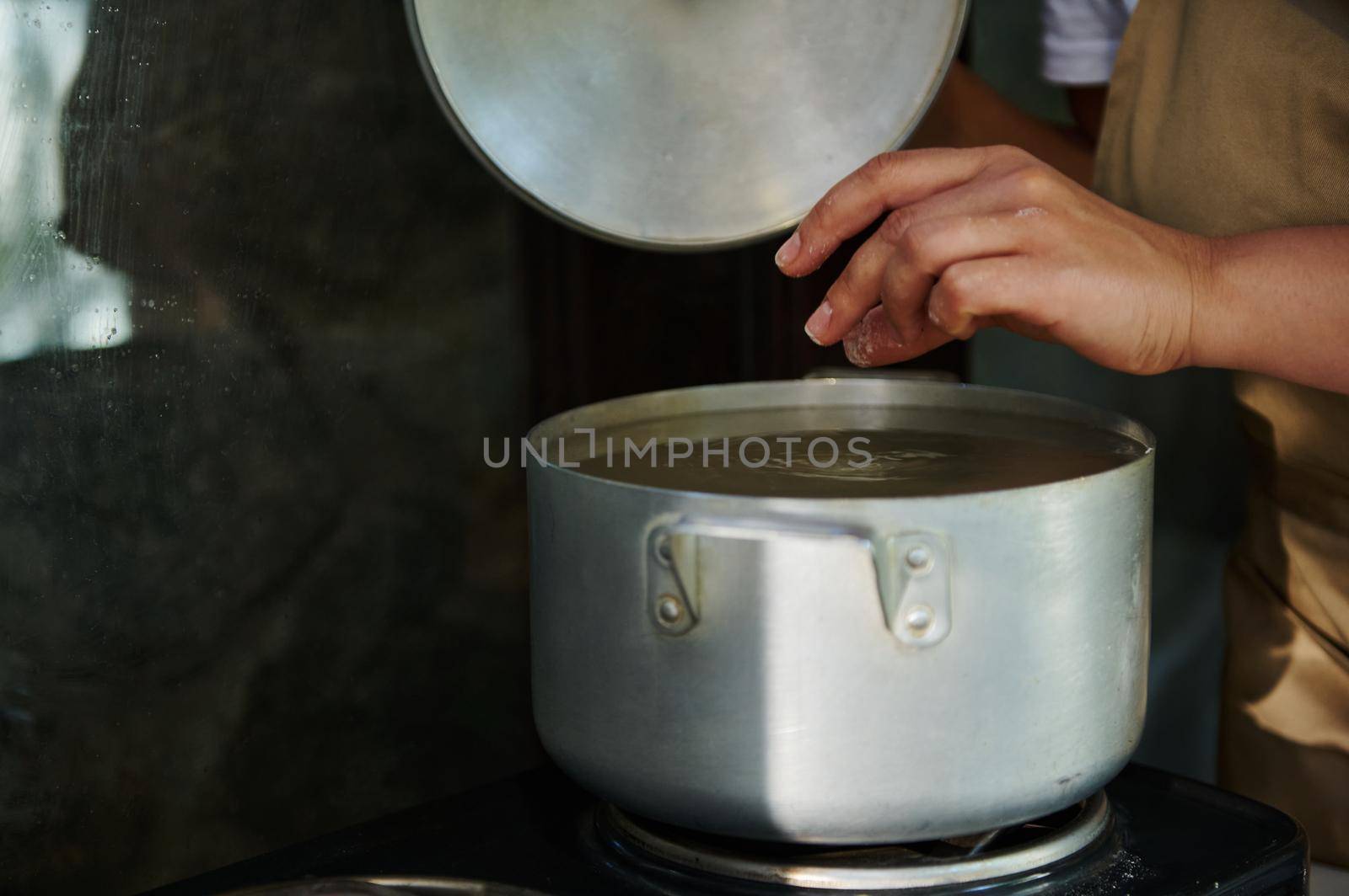 Close-up. Cropped view of a housewife standing by a kitchen table with electric stove, opening the lid of a metal saucepan, dipping raw dumplings into a boiling water, brewing Ukrainian vareniki.