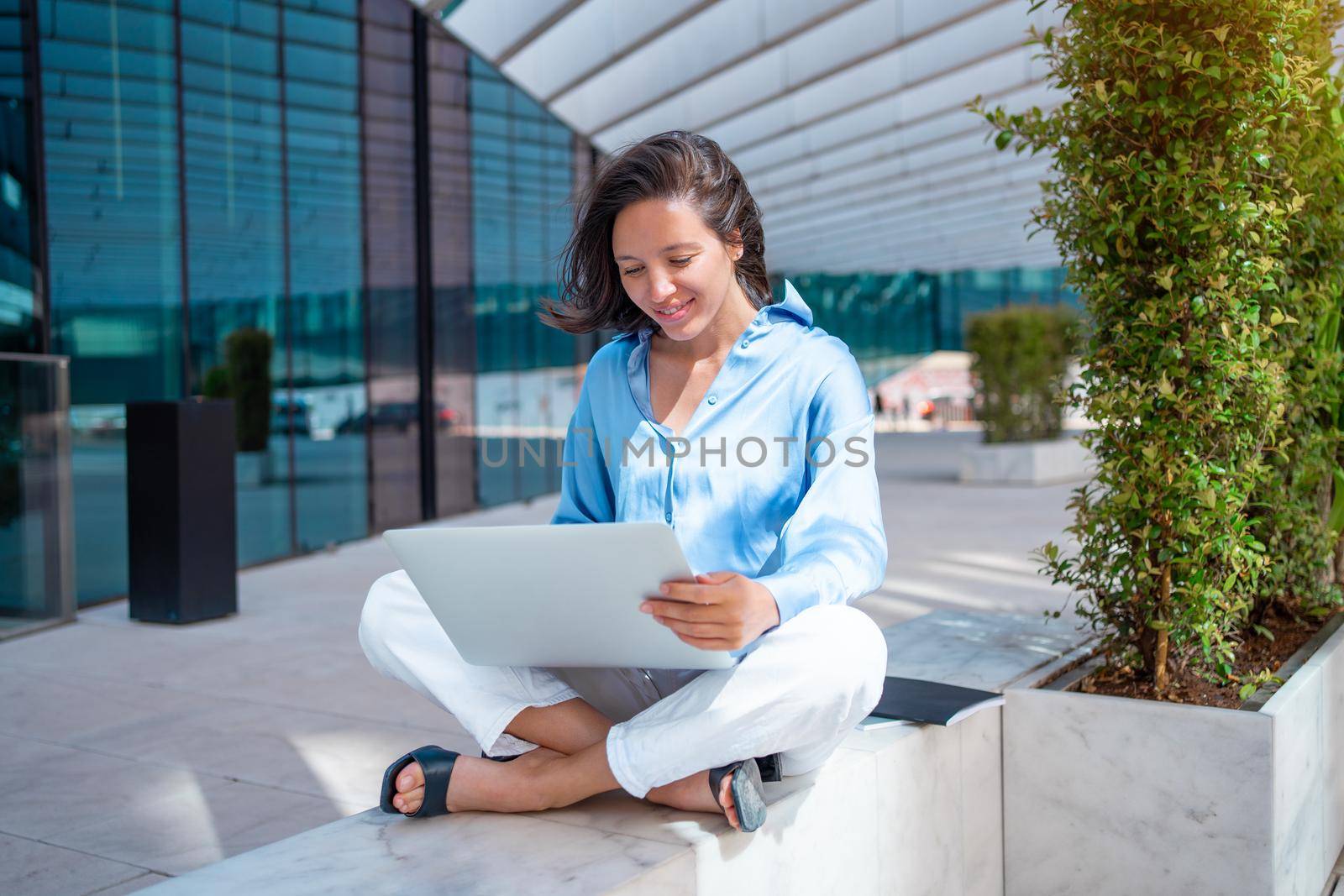 Pretty business woman use laptop outside sitting lotus position near office building. Positive smile caucasian female business person do remote work outdoors. Self employed freelance worker