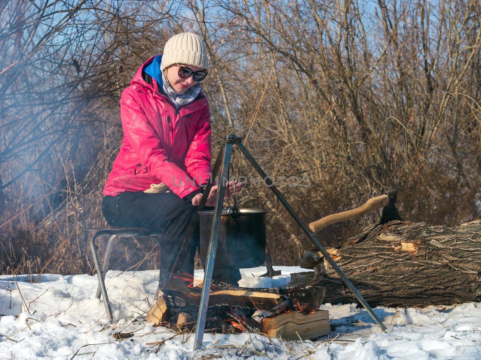 Female caucasian in pink jacket with sunglasses warms her hands by campfire, pot of soot over bonfire hanging on tripod, ax in tree trunk on backdrop, winter outdoor cooking at campsite, lifestyle