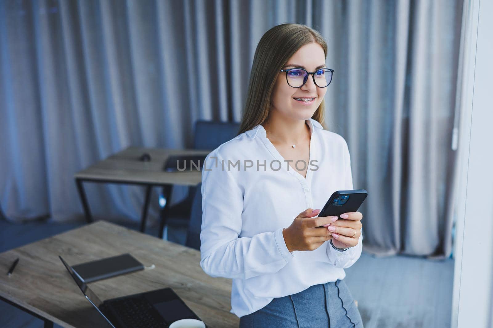 Photo of a successful young woman in a shirt smiling and working on a laptop while talking on the phone in a modern office with large windows. Remote work with coffee