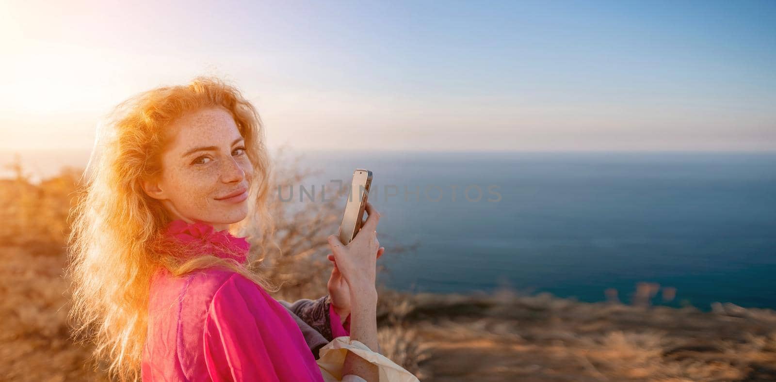 Close up shot of beautiful young caucasian woman with curly blond hair and freckles looking at camera and smiling. Cute woman portrait in a pink long dress posing on a volcanic rock high above the sea