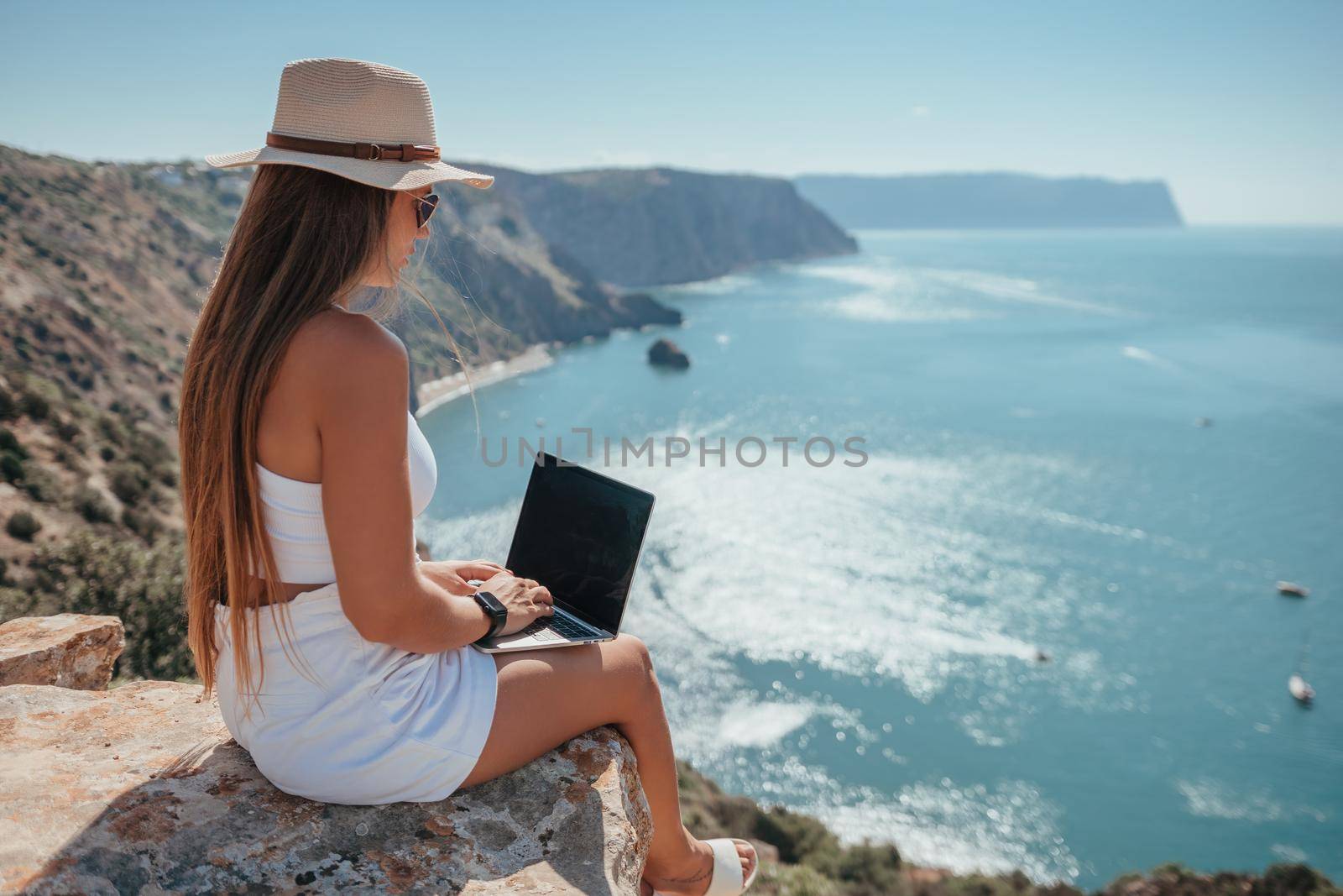 Successful business woman in yellow hat working on laptop by the sea. Pretty lady typing on computer at summer day outdoors. Freelance, travel and holidays concept.