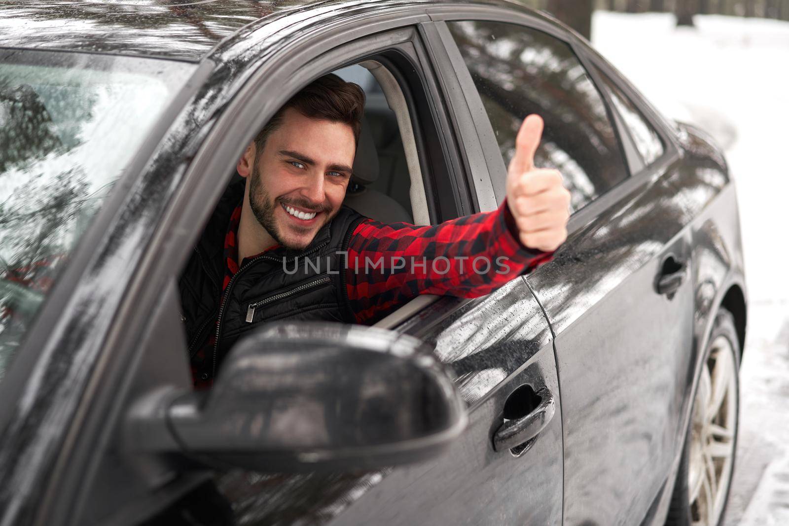 Young adult attractive Caucasian man sits at the wheel of his car sunny winter day shoeing thumbs up gesture. Wintertime road trip. Happy smiling hipster guy sitting in car and looking window.
