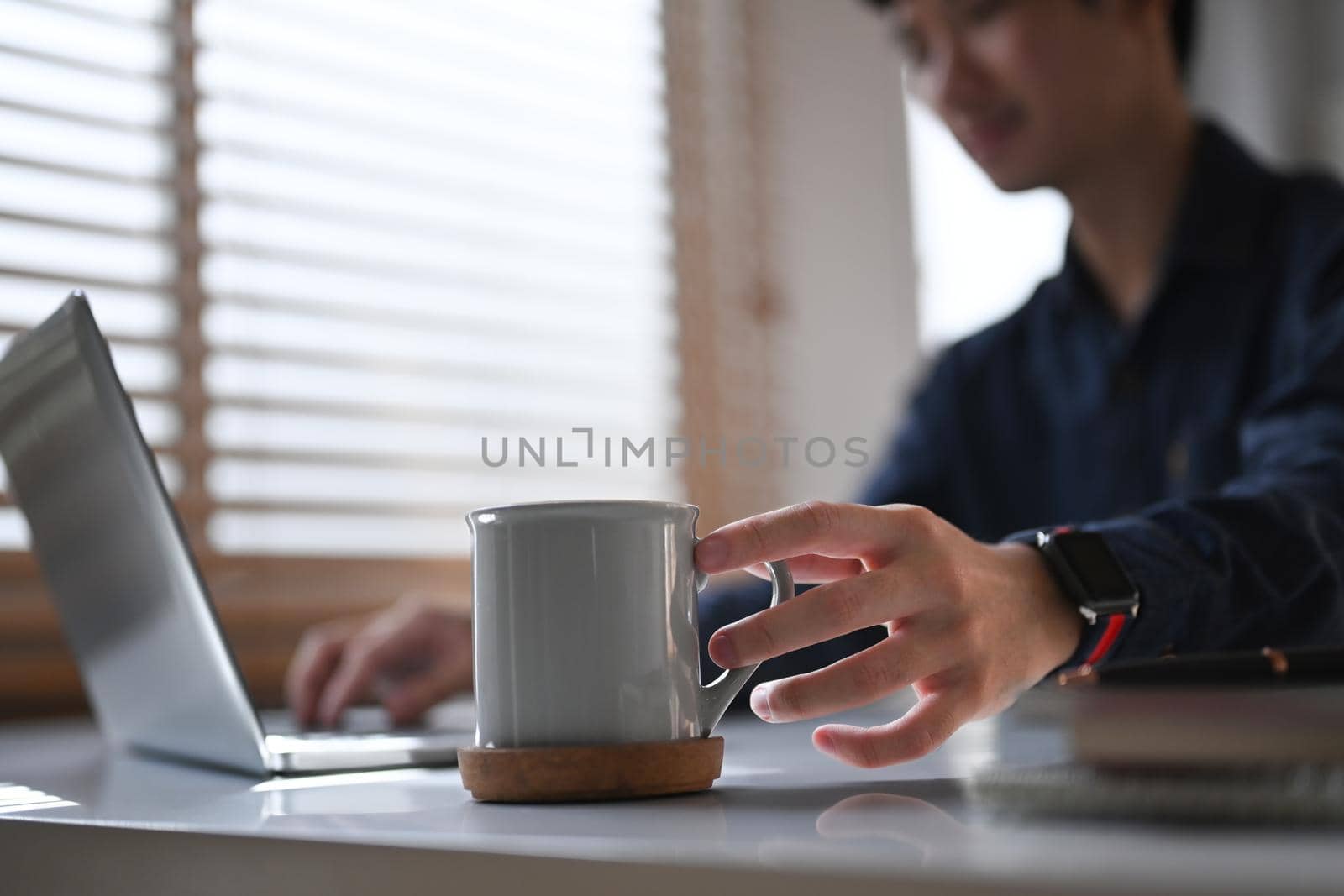 Close up view businessman hands holding cup of coffee and using laptop computer at office desk by prathanchorruangsak