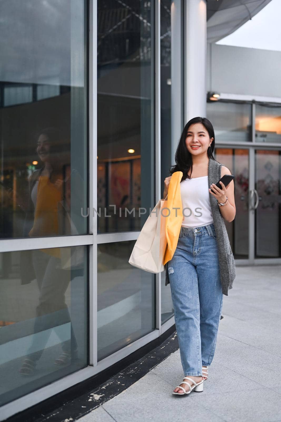 Stylish young woman with shopping bags walking outside shopping mall in the evening.