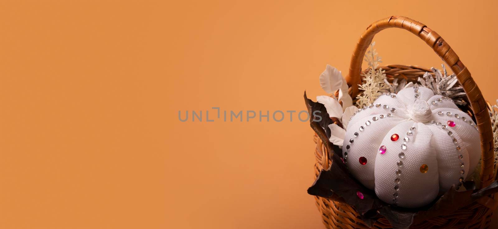 Banner with white decorative hand made pumpkin with shiny stones and fall leaves in basket on colored background. Autumn harvest concept.