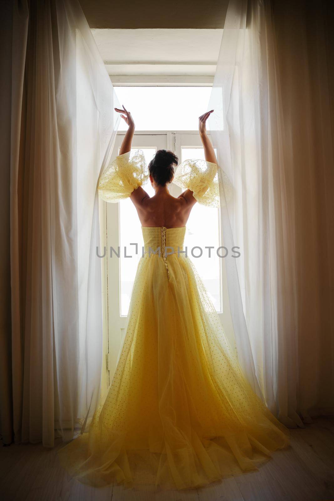 A woman's silhouette in a golden luxurious dress against the background of a window holds a curtain with her hands. Elegant lady in a yellow long silk dress with bare back, back view. by Matiunina