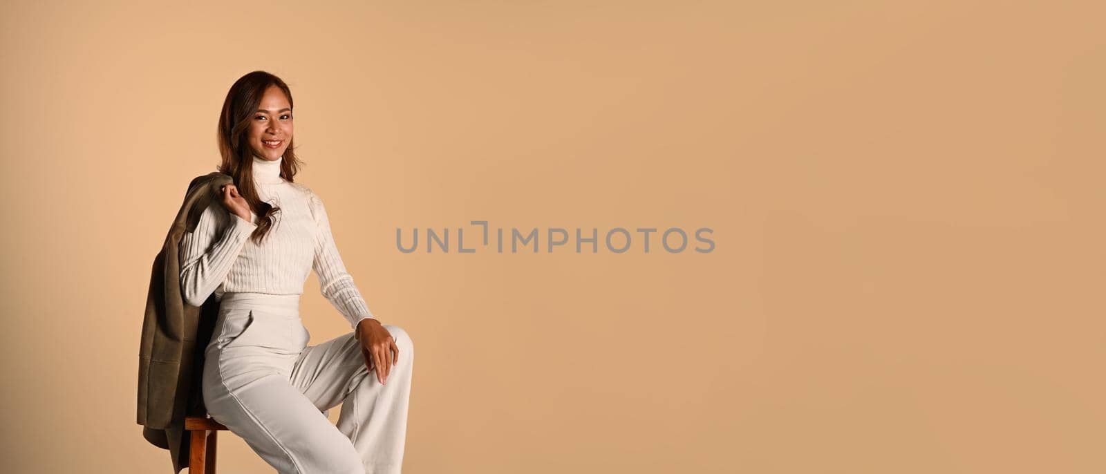 Stylish young woman with trench coat on shoulder looking at camera, sitting on beige background with copy space for your advertise text.
