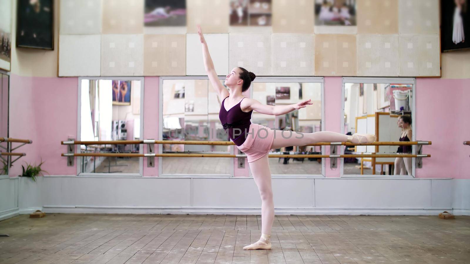 in dancing hall, Young ballerina in black leotard performs 1 arabesque, raises her leg up behind elegantly, standing near mirror in ballet class. High quality photo