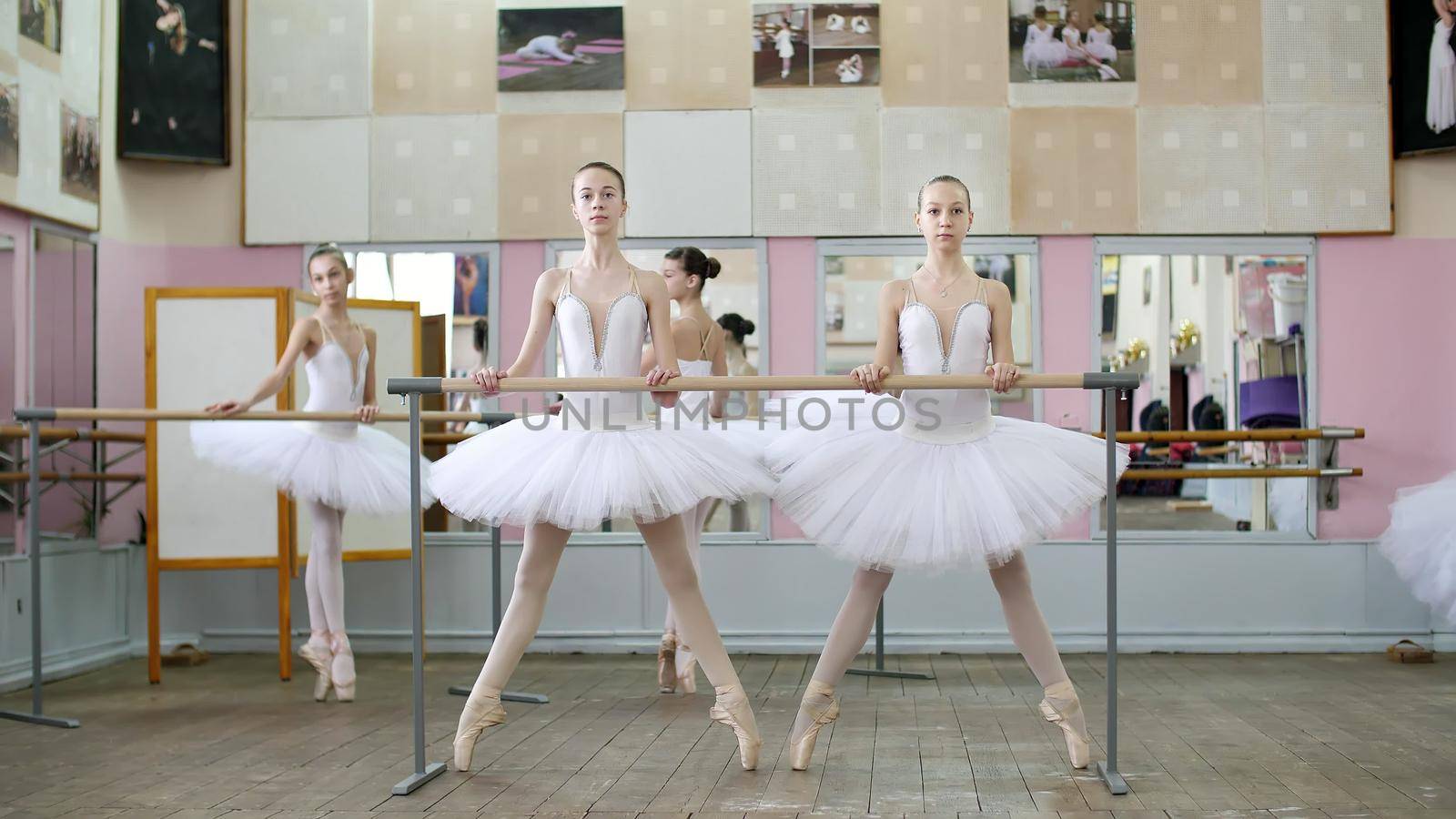 in the ballet hall, girls in white ballet tutus, packs are engaged at ballet, rehearse plie forward, Young ballerinas crouch standing on toes in pointe shoes at railing in ballet hall. High quality photo