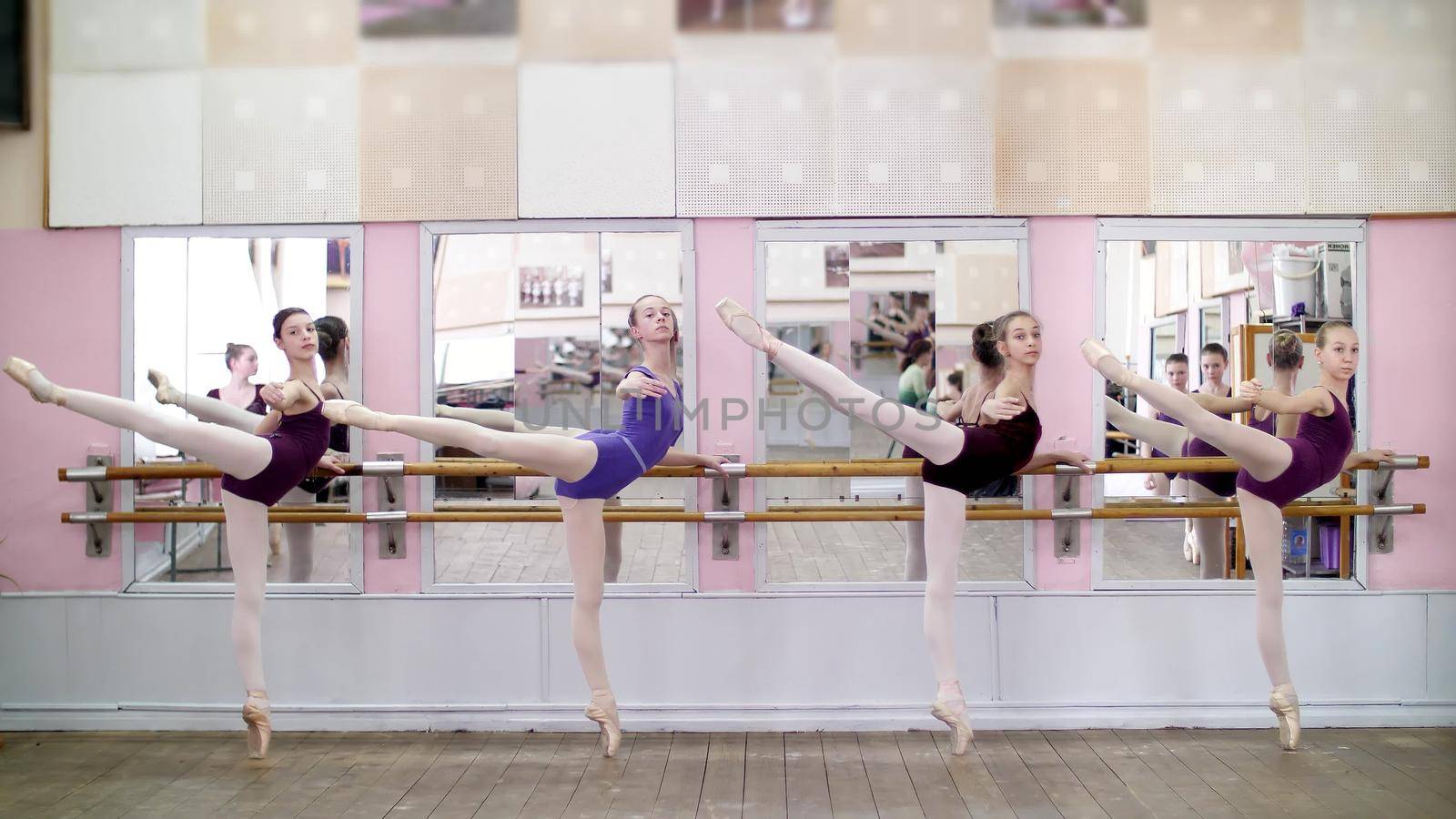 in dancing hall, Young ballerinas in purple leotards perform attitude efface on pointe shoes, standing near barre at mirror in ballet class. High quality photo