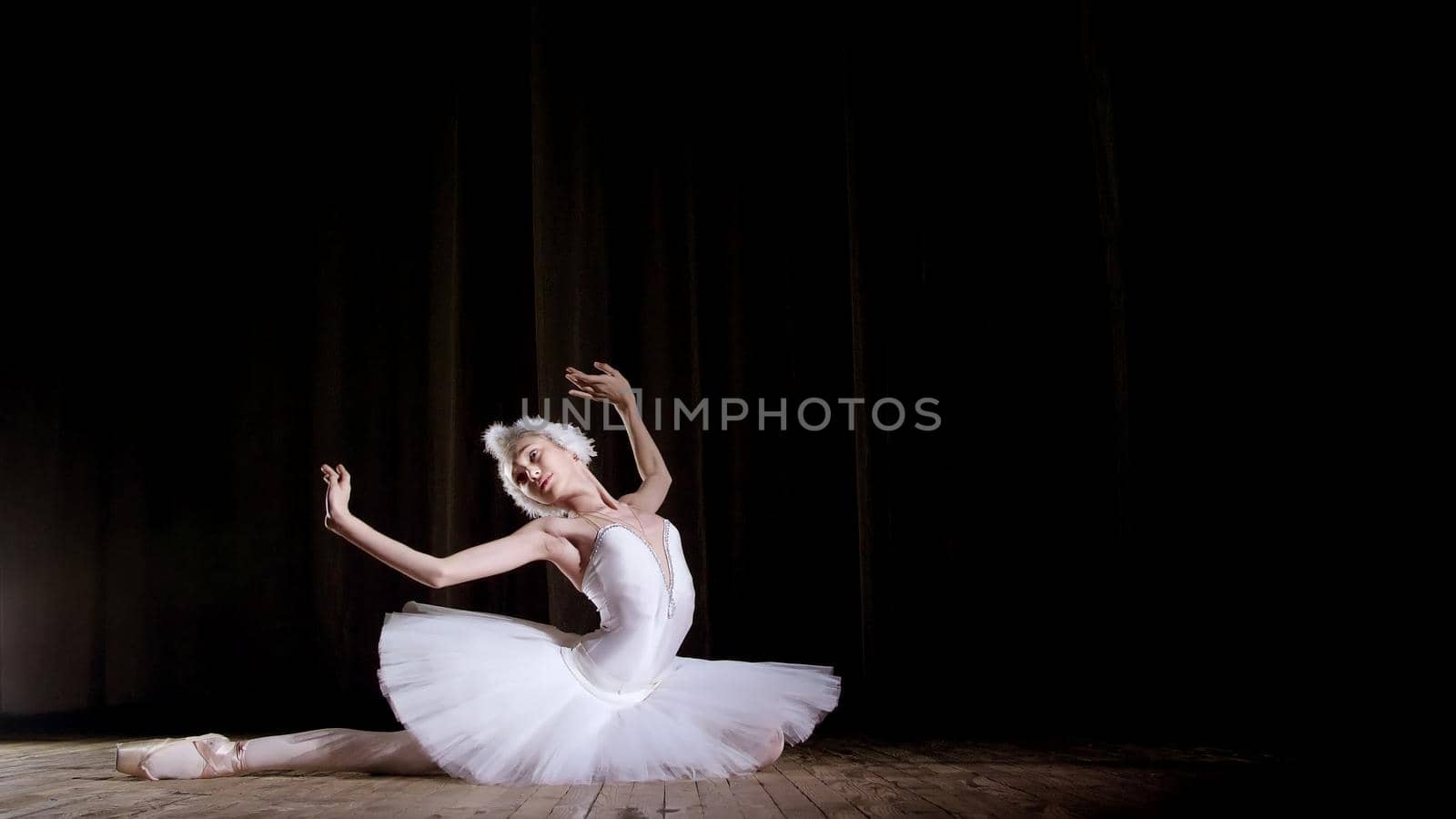 in rays of spotlight, on the stage of the old theater hall. Young ballerina in suit of white swan and pointe shoes, dances elegantly certain ballet motion, Swan Lake. High quality photo