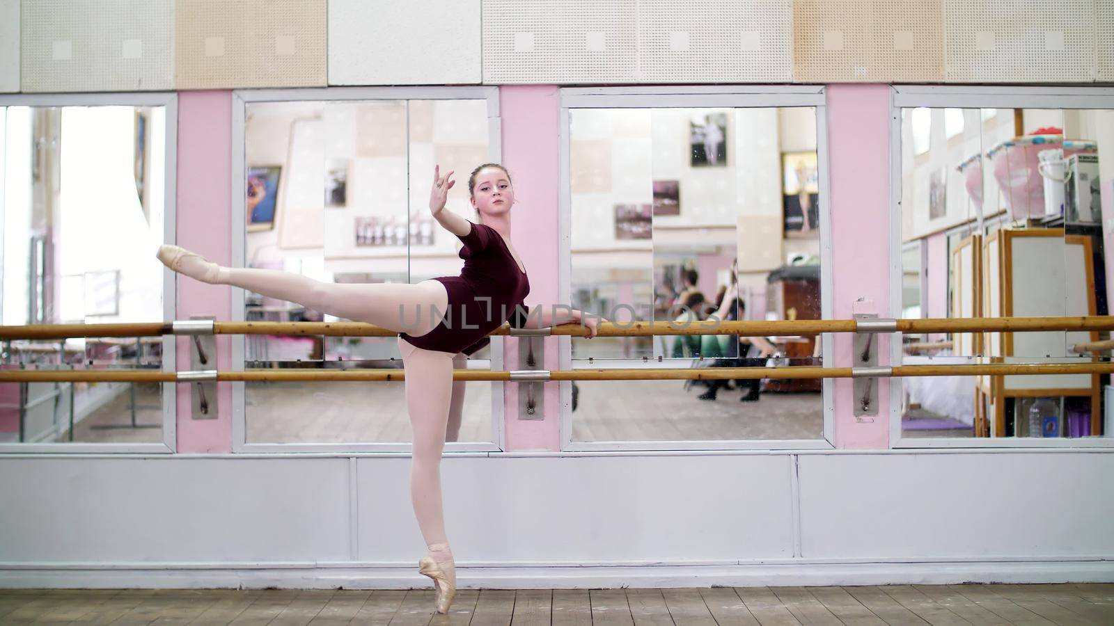 in dancing hall, Young ballerina in purple leotard performs developpe attitude on pointe shoes, raises her leg up behind elegantly, standing near barre at mirror in ballet class. High quality photo