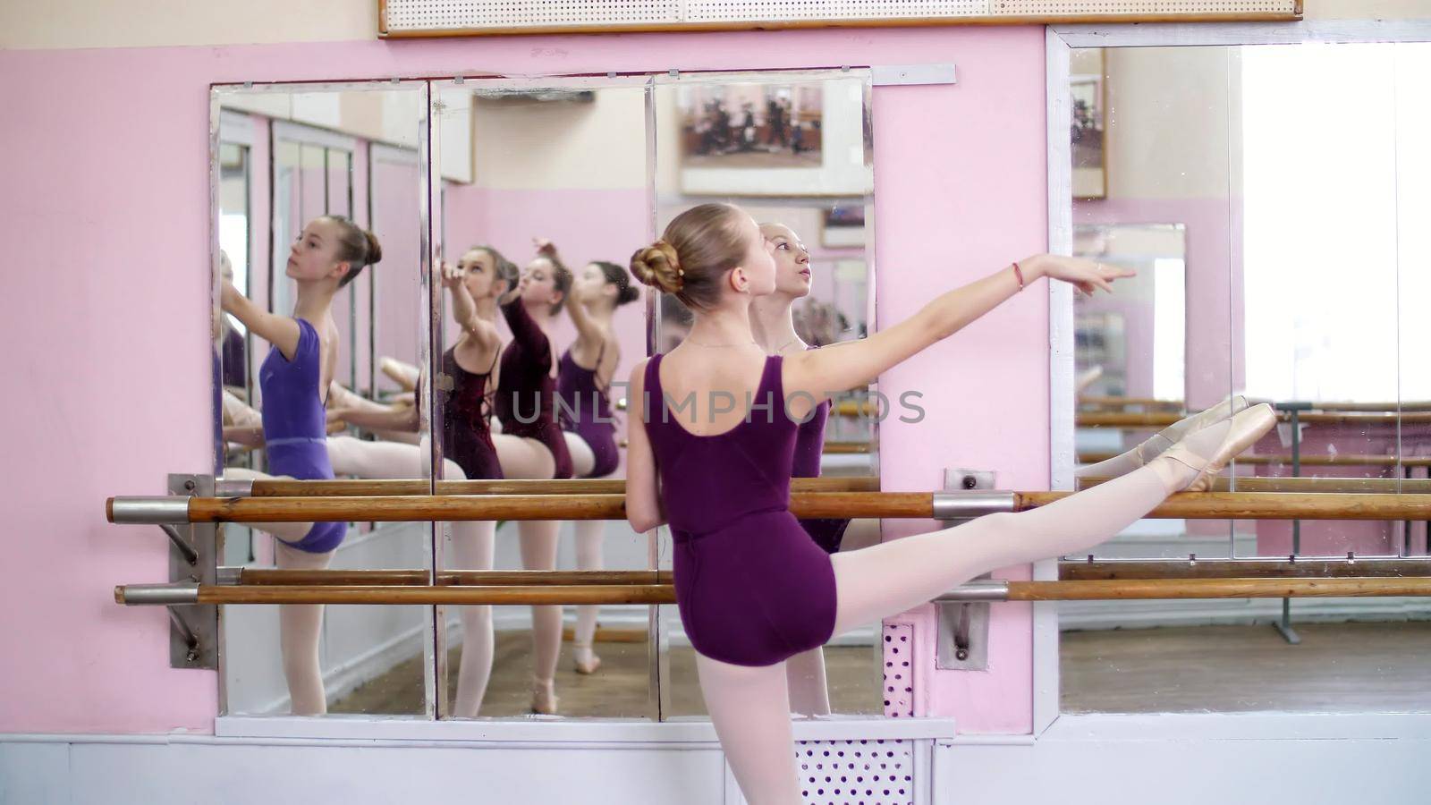 Girl standing at railing in ballet hall. Young ballerina stretching out using ballet barre in studio. Young ballerina in purple leotard. High quality photo