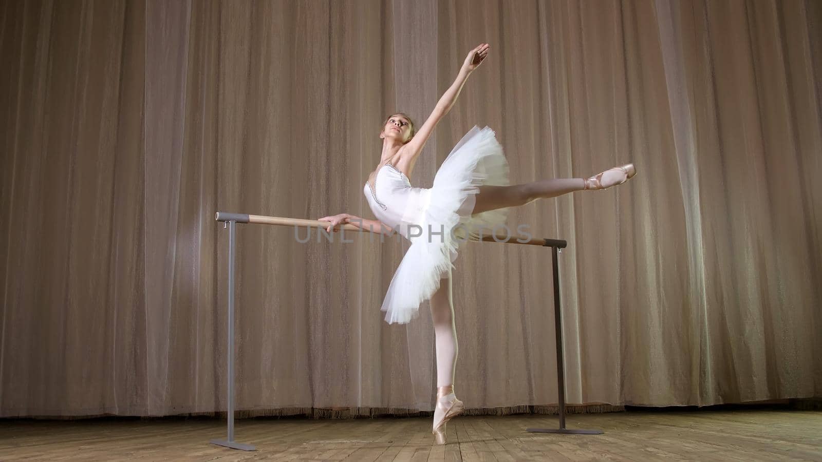 ballet rehearsal, in old theater hall. Young ballerina in white ballet skirt, tutu, is engaged at ballet, performs elegantly a certain ballet exercise, attitude allonge, standing near barre. High quality photo
