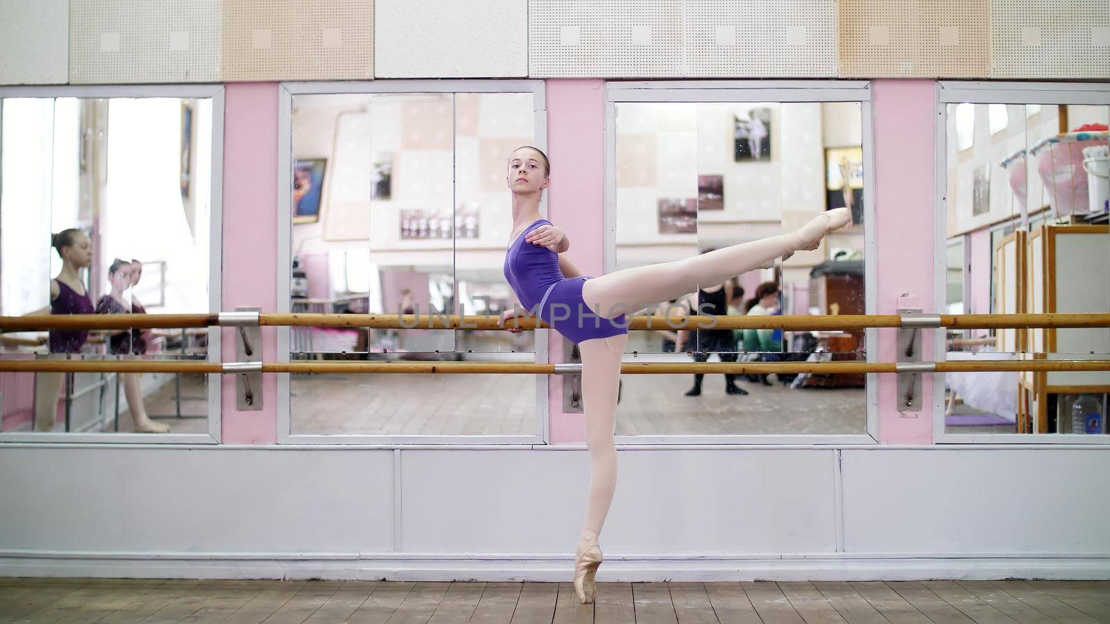 in dancing hall, Young ballerina in purple leotard performs developpe attitude on pointe shoes, raises her leg up behind elegantly, standing near barre at mirror in ballet class. High quality photo