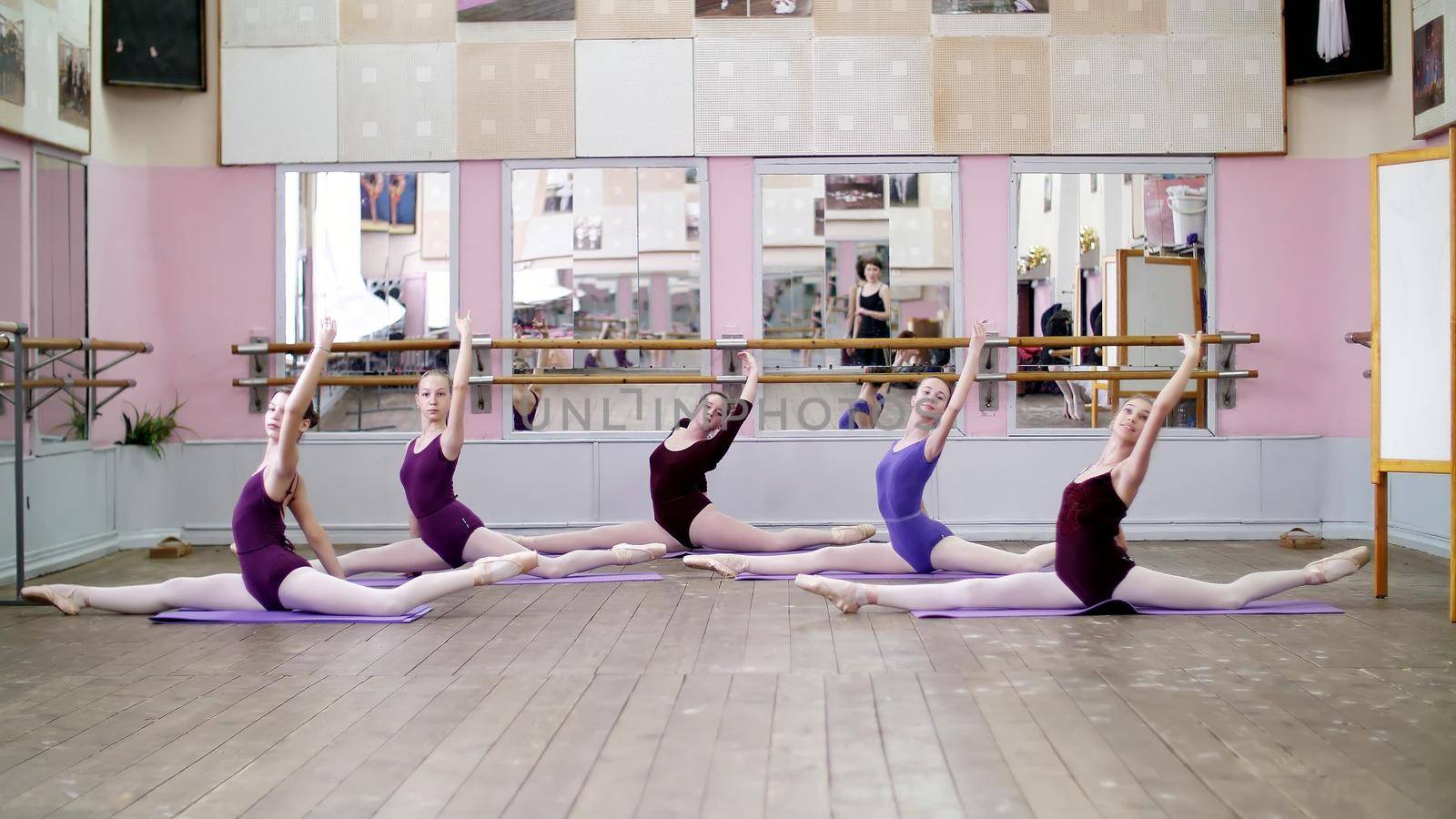 in dancing hall, Young ballerinas in purple leotards perform part de bras In 3 position with forward tilt, girls are sitting on twines, raise their legs up behind elegantly, at mirror in class. by djtreneryay