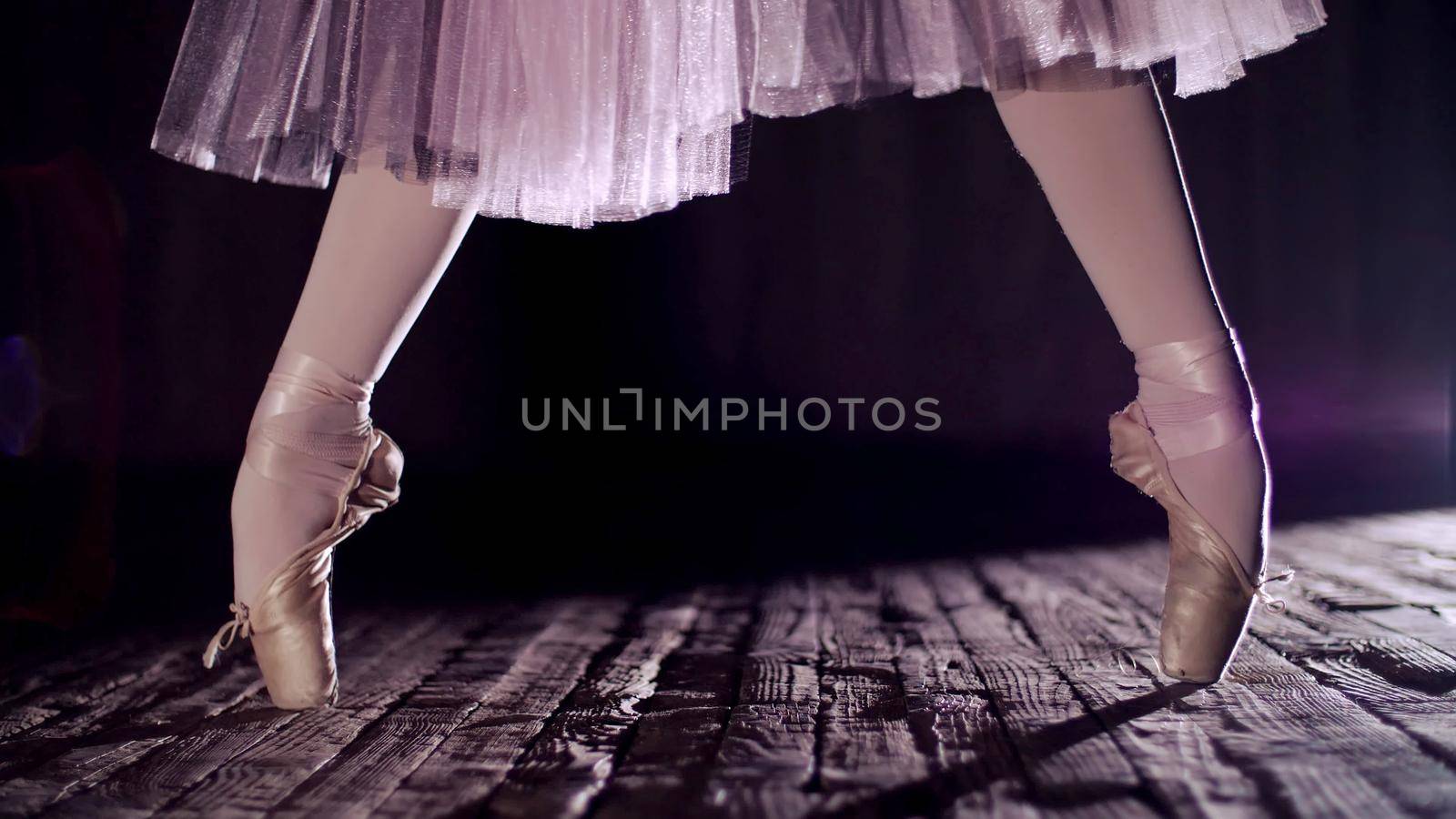 close up, in rays of spotlight, on the stage of the old theater hall. ballerina in white ballet skirt, raises on toes in pointe shoes, performs elegantly a certain ballet exercise, relleve. High quality photo