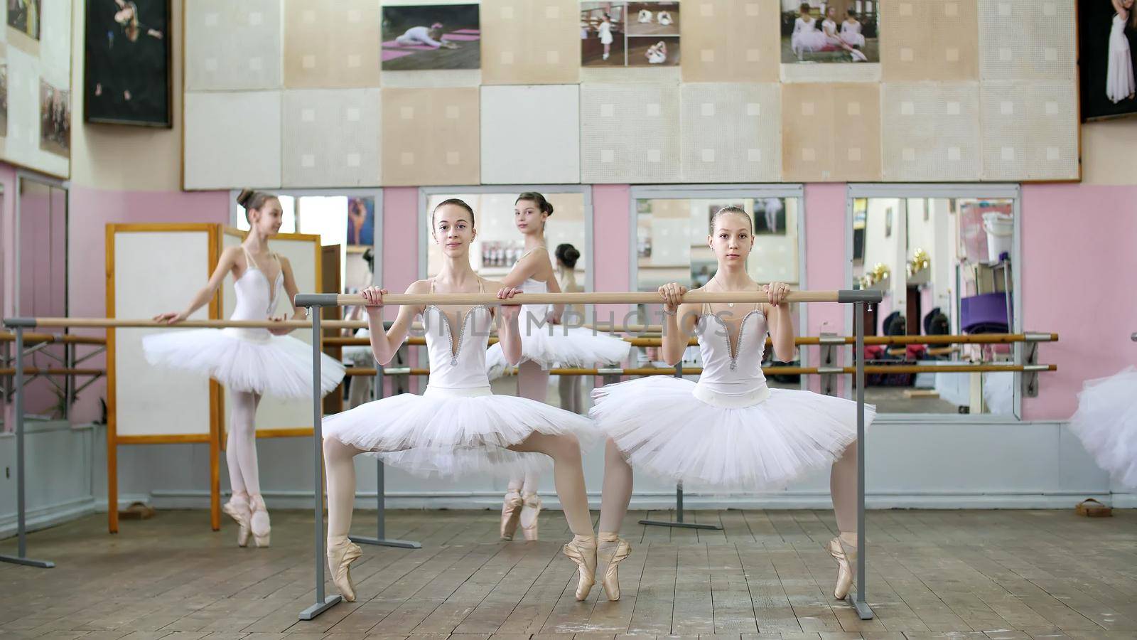 in the ballet hall, girls in white ballet tutus, packs are engaged at ballet, rehearse plie forward, Young ballerinas crouch standing on toes in pointe shoes at railing in ballet hall. by djtreneryay