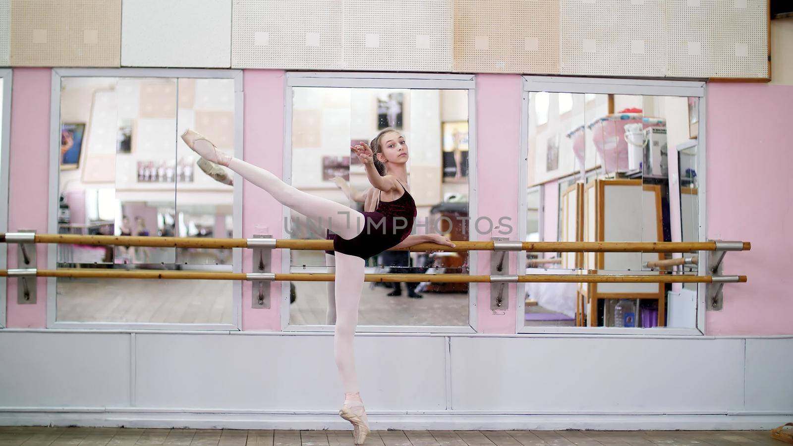 in dancing hall, Young ballerina in purple leotard performs developpe attitude on pointe shoes, raises her leg up behind elegantly, standing near barre at mirror in ballet class. High quality photo