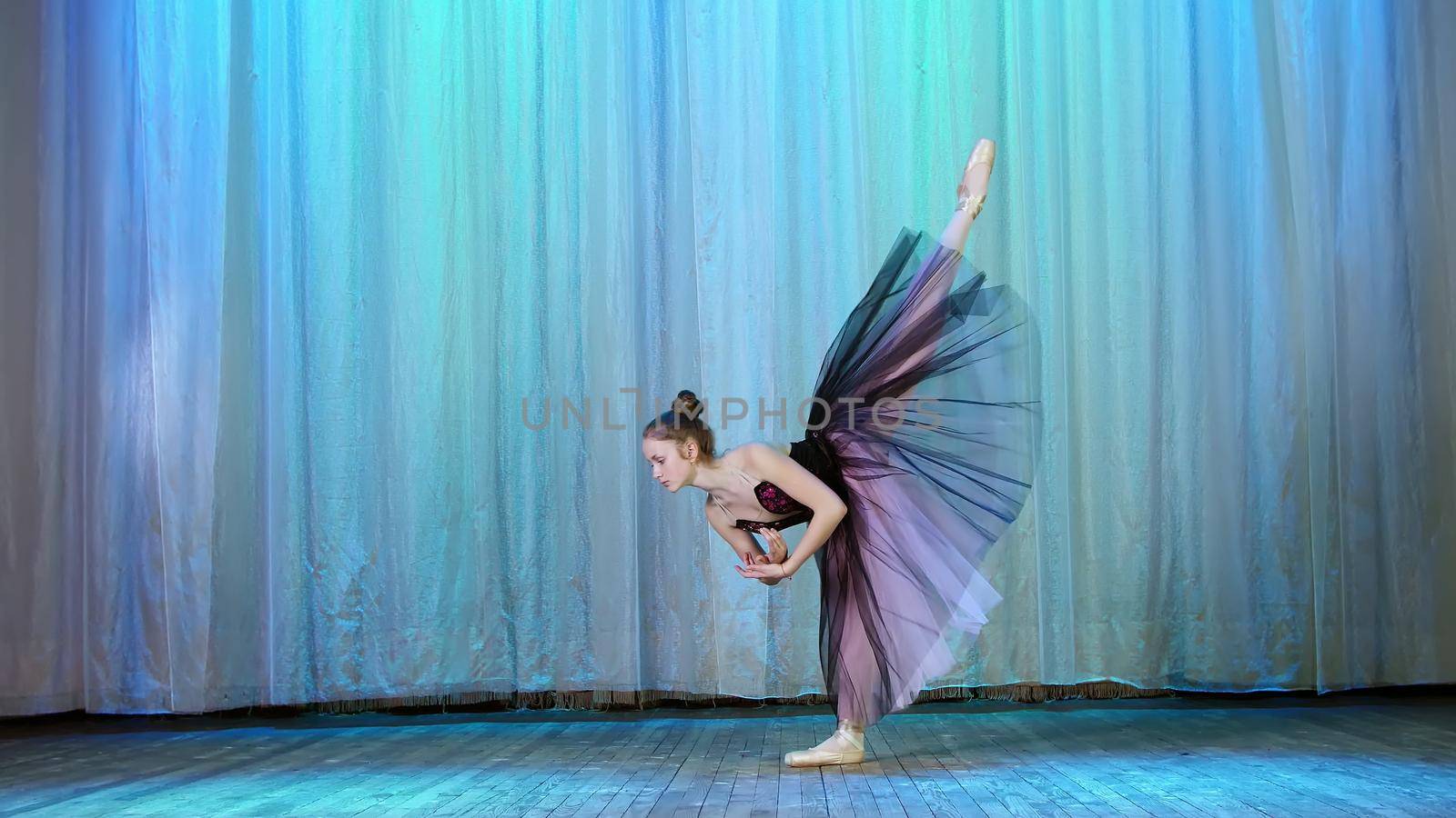 ballet rehearsal, on the stage of the old theater hall. Young ballerina in lilac black dress and pointe shoes, dances elegantly certain ballet motion, raises her leg up behind. High quality photo