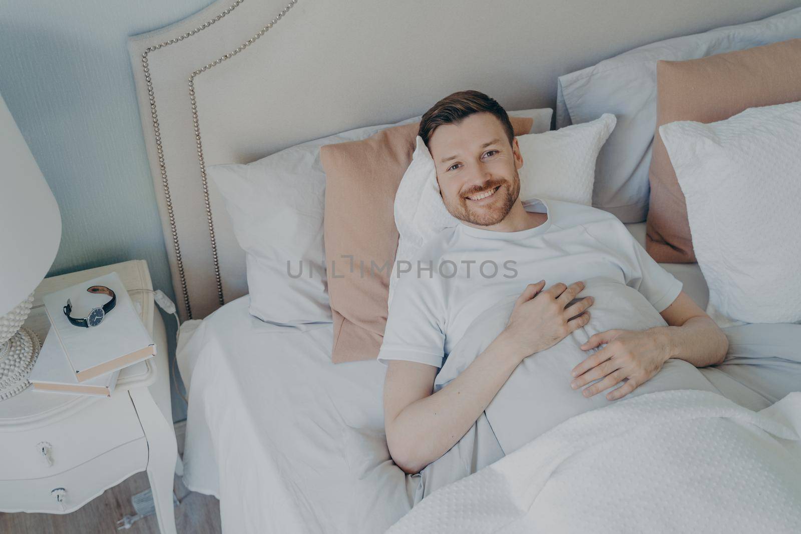 Young well rested man in white tee shirt waking up in morning with smile, happy after sleeping enough, lying in comfortable bed while covered with blankets, lots of different pillows