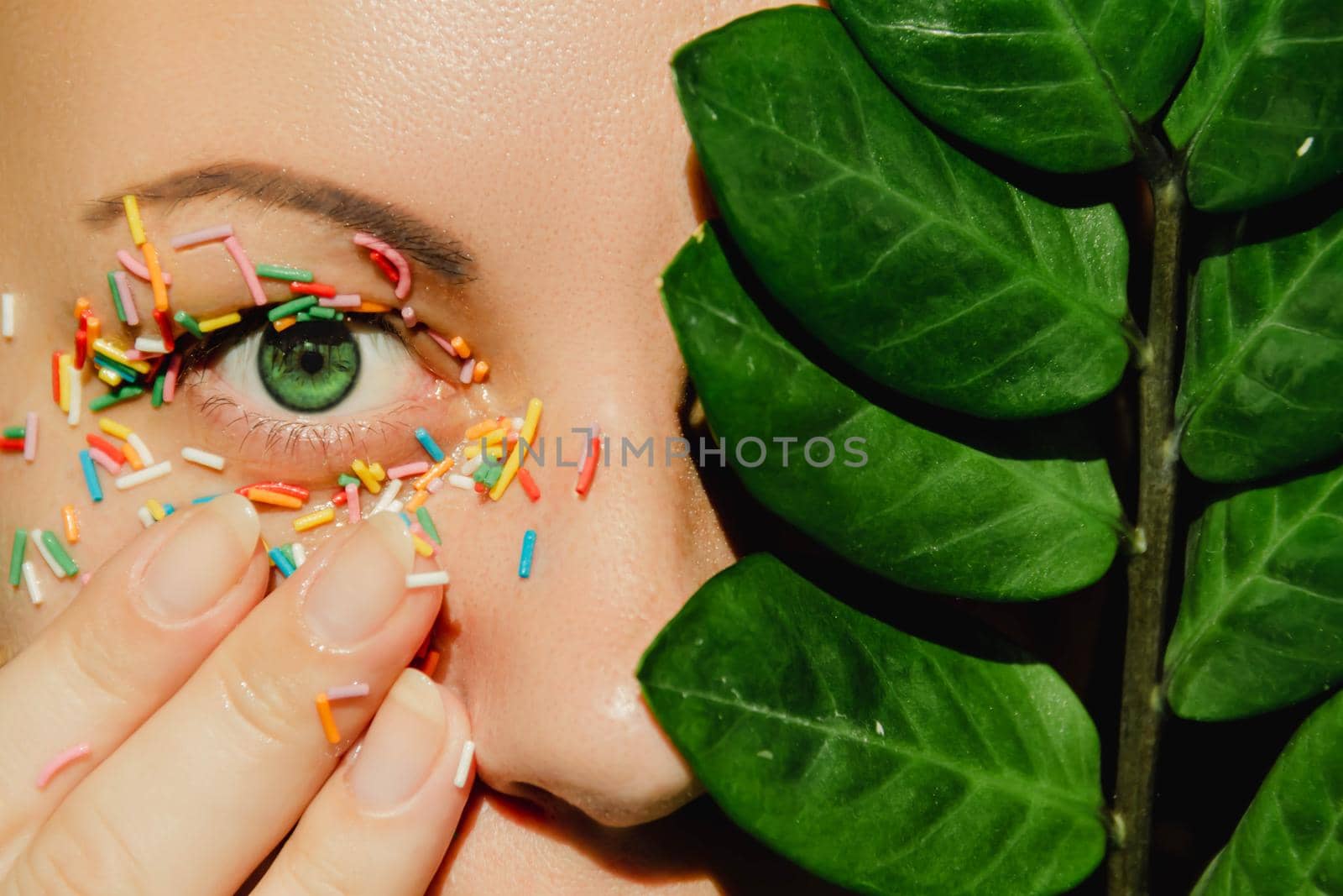 An open, green woman's eye with a sweet, multicolored sprinkle on the eyelid and a plant with large, green leaves