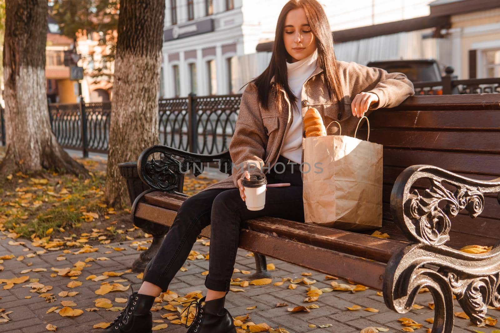 A stylish brunette woman walks around the autumn city. The brunette is sitting on a bench outside with a glass of coffee and a phone, holding a craft bag with a baguette.