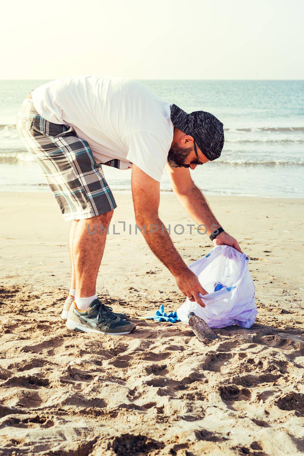 vertical photo of an activist picking up plastics and trash that pollutes the sea on a bag on a sunny day, cleaning on the beach, campaign to clean volunteer concept