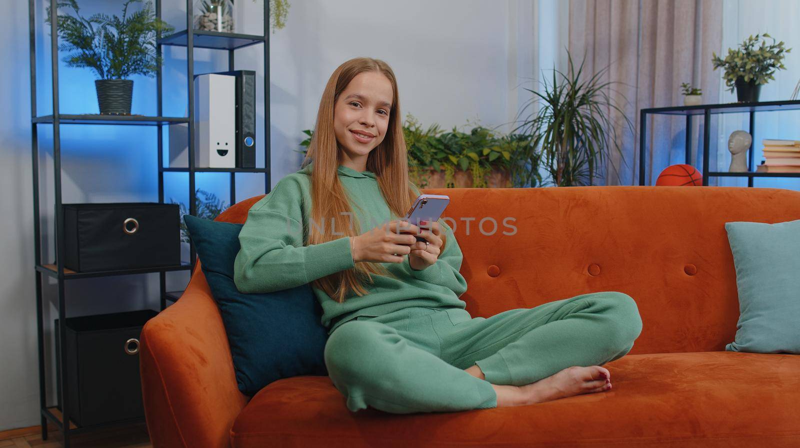 Portrait of girl sitting on sofa uses mobile phone smile at modern home apartment. Young child kid texting share messages content on smartphone social media applications online, watching relax movie