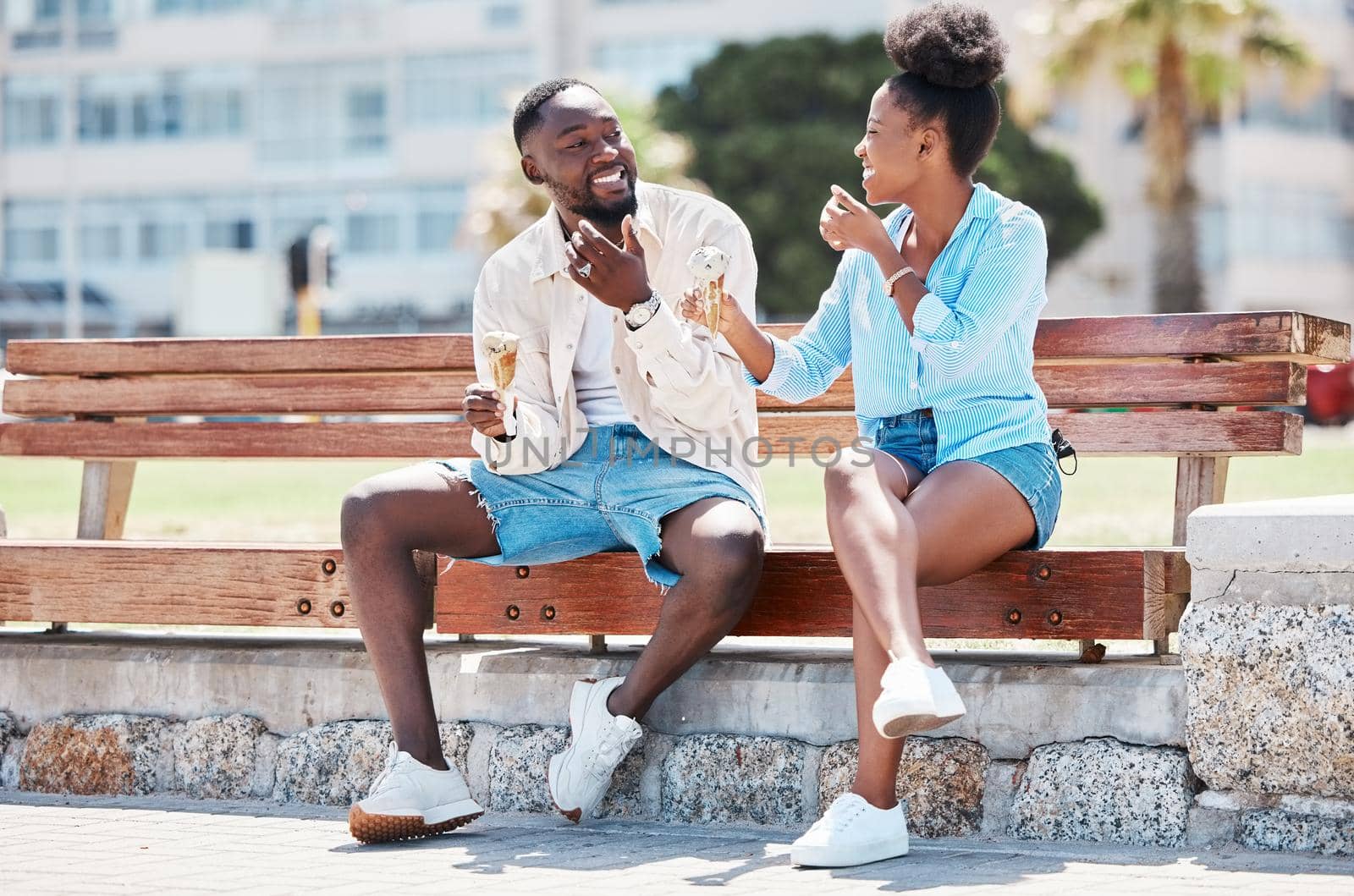 Happy black couple eating ice cream on a beach bench together, smiling while bonding and laughing. Young African American man and woman enjoying their summer romance, free time and relationship.