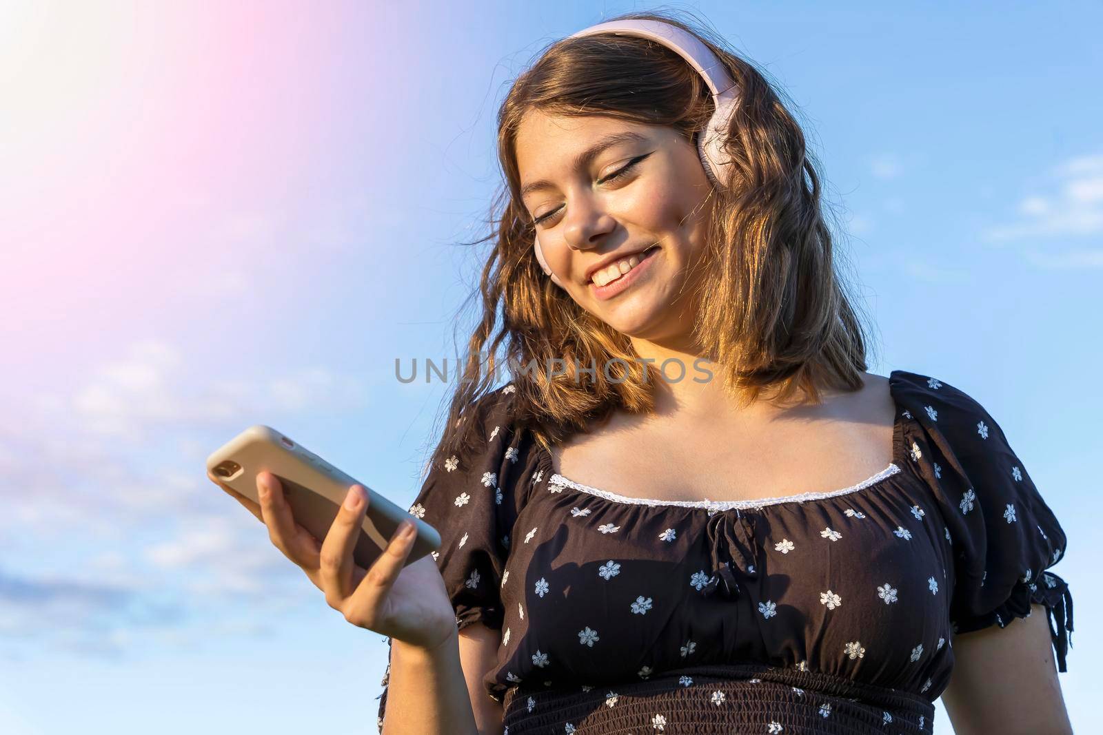 A contented smiling brunette in a dress listens to music on her smartphone through wireless headphones against a blue sky background