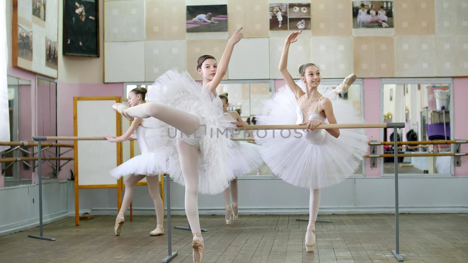 in ballet hall, girls in white ballet skirts are engaged at ballet, rehearse attitude, Young ballerinas standing on toes in pointe shoes, raise legs up behind elegantly, at railing in ballet hall. by djtreneryay