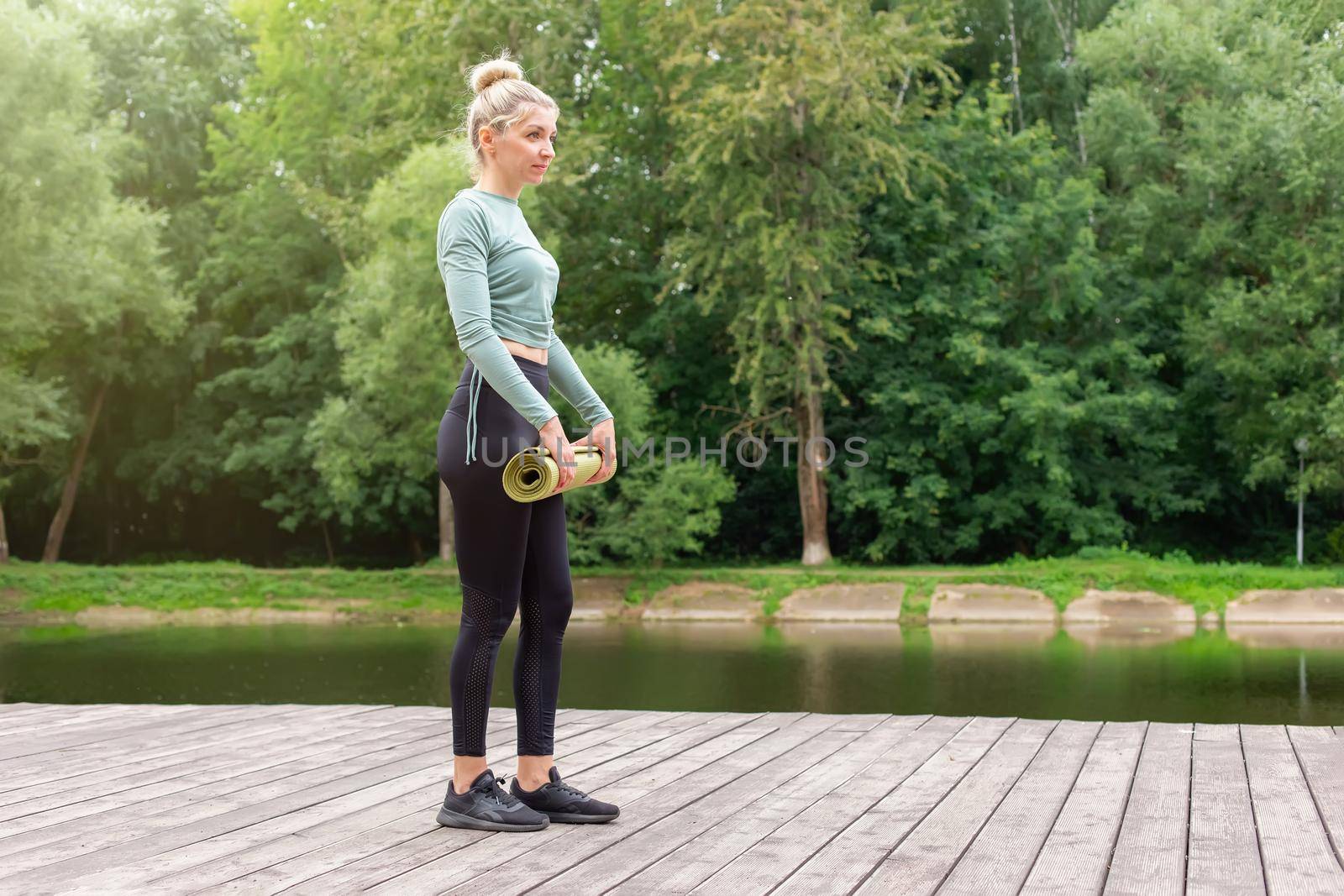 A blonde woman in a green top and black leggings, standing in the park by pond in summer, holding a green gym mat, preparing for sports. the side view. Copy space