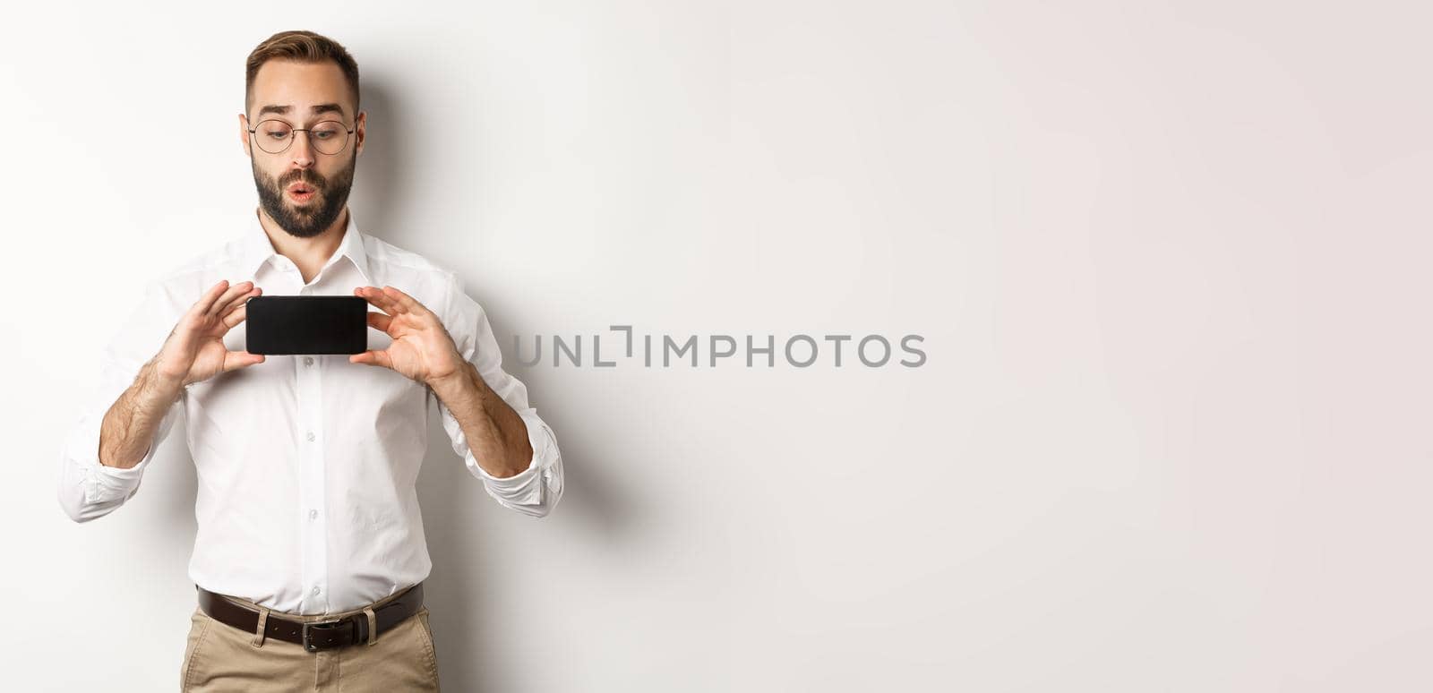 Amused handsome guy showing mobile screen, looking excited at online website, standing over white background.