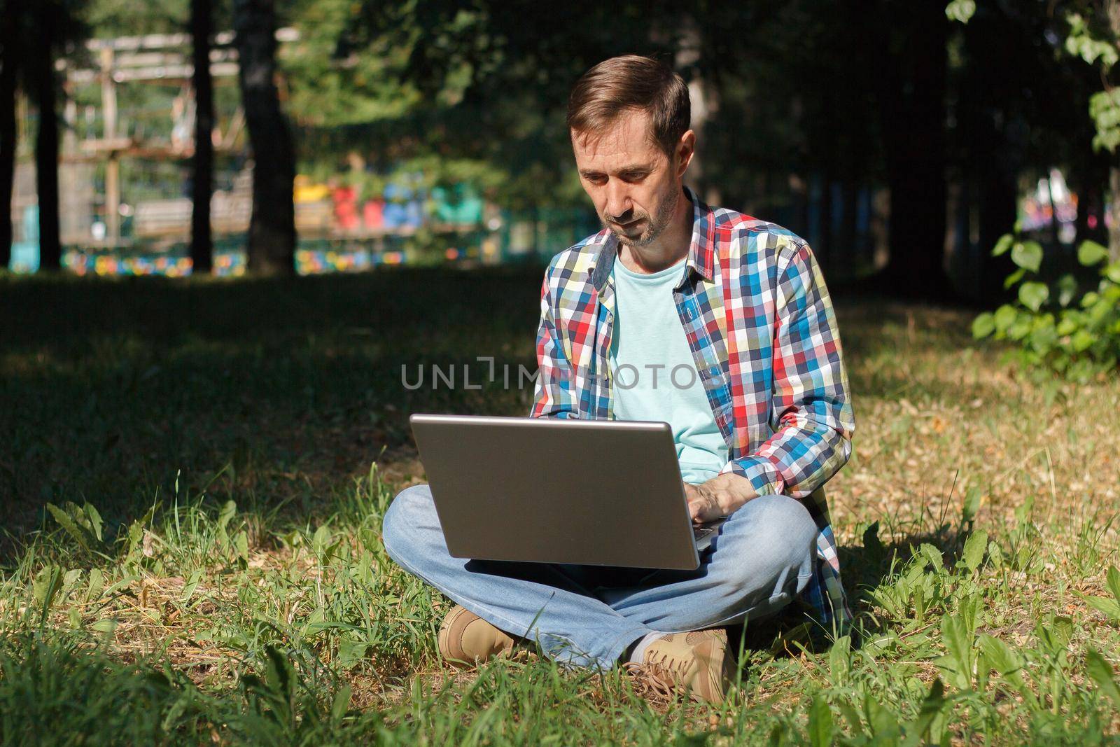 Adult man works on his computer in the park on the lawn.Remote work concept. The writer works remotely, enjoying nature.