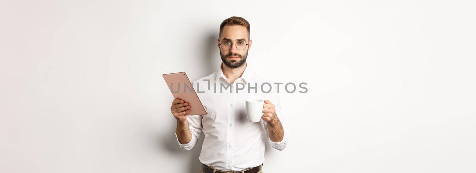 Confident male manager reading work on digital tablet and drinking coffee, standing over white background.