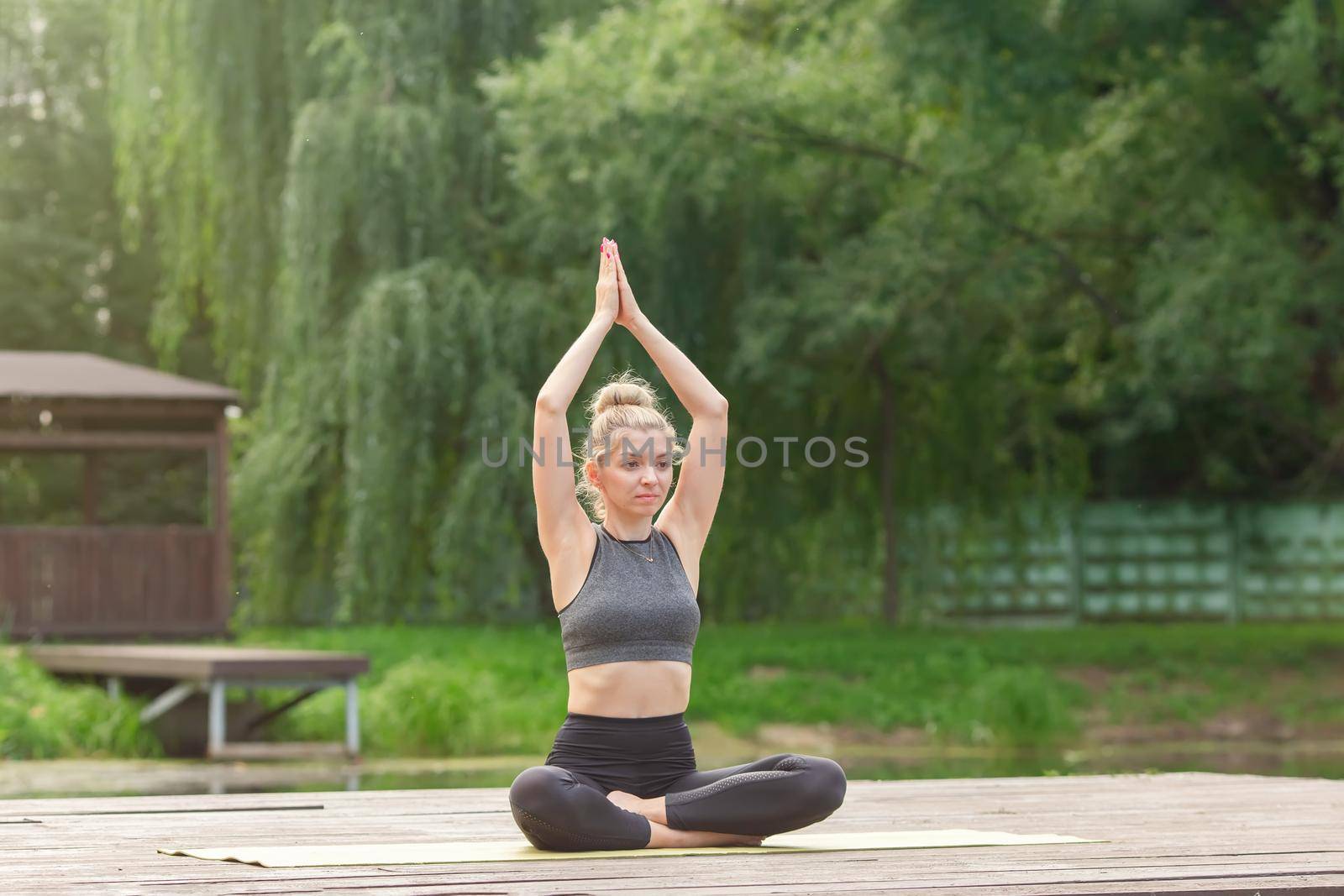 A slender woman in a gray top and leggings, sitting on a wooden platform by a pond in the park in summer, does yoga with her closed palms raised above her head. Copy space