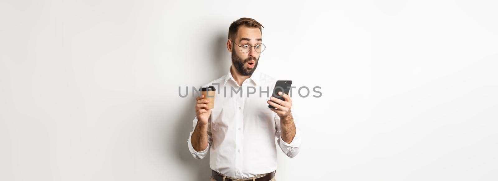 Businessman drinking coffee and looking surprised at message on mobile phone, standing amazed over white background.