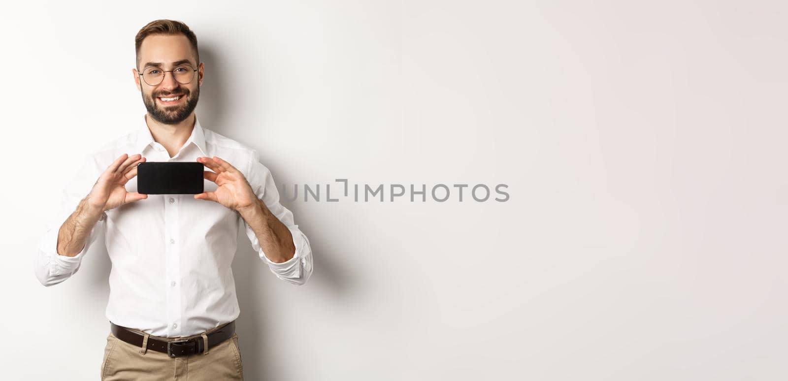 Handsome bearded man in glasses, demonstrating mobile phone application, showing smartphone screen, standing over white background.