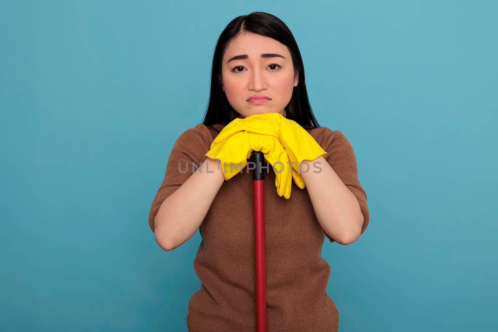 Overwhelmed sad and tired asian houseworker taking some rest wearing yellow gloves holding on brush stick isolated against a blue background, Cleaning home concept, Overworked woman feeling negative