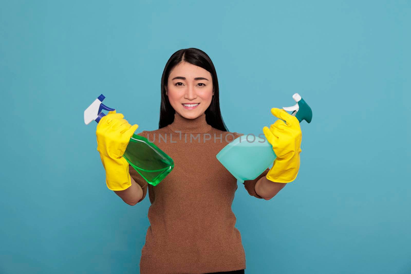 Cheerful joyful asian woman holding two detergent spray in yellow gloves isolated on a blue background, Cleaning home concept, smiling happy housewife worker female at chores