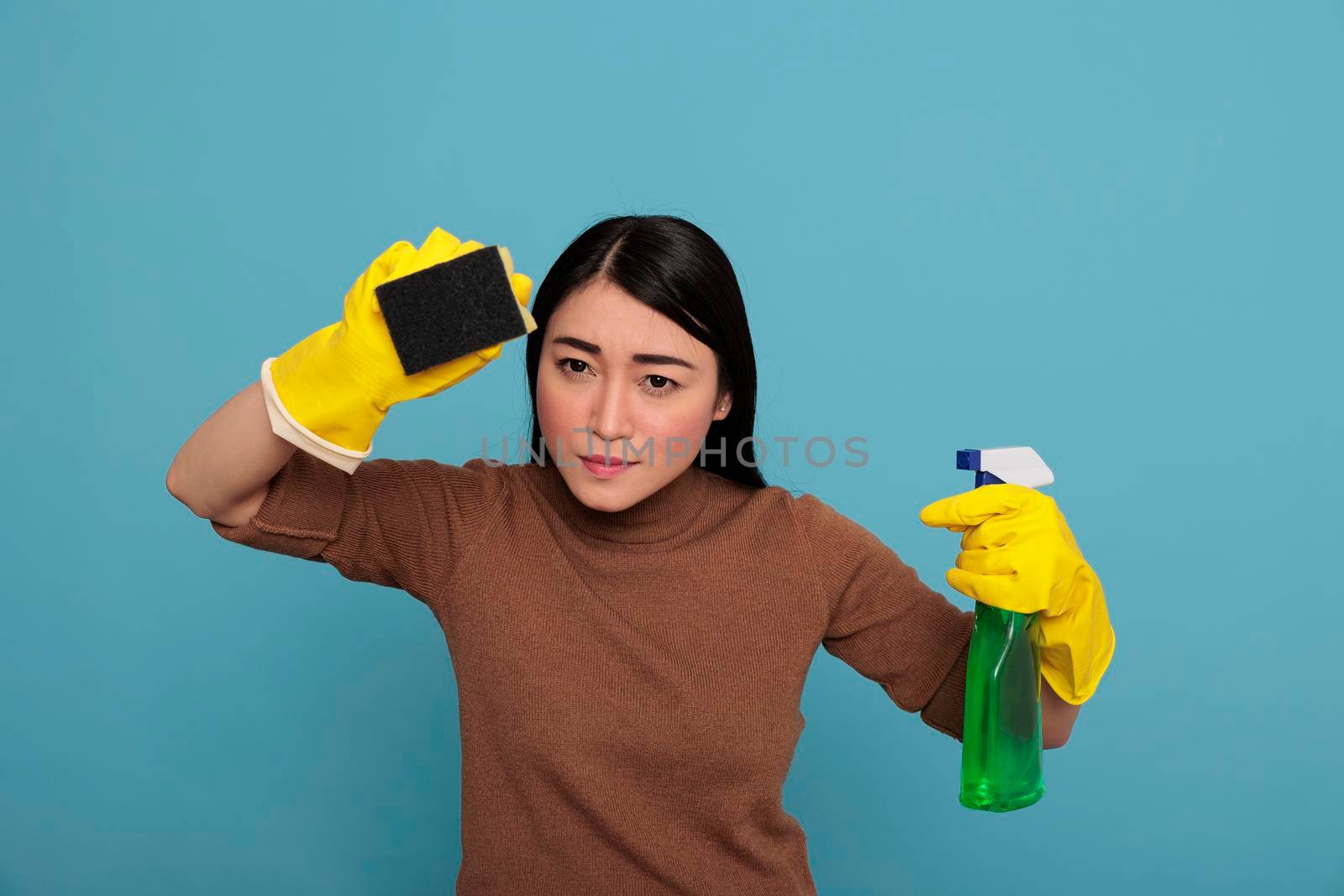 Portrait of serious young woman wearing yellow gloves holding a sponge detergent sprayer isolated on a blue background, Housewife worker, Cleaning home concept, Female angry face expression