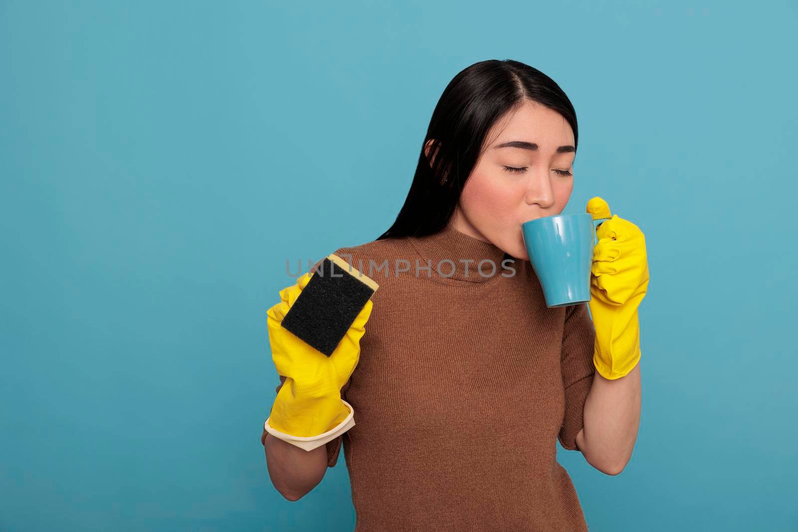 Asian tired young asian housewife taking coffee during work break while holding a sponge in the other hand, Cleaning home concept, Refreshment tea take some rest from day to day chores,