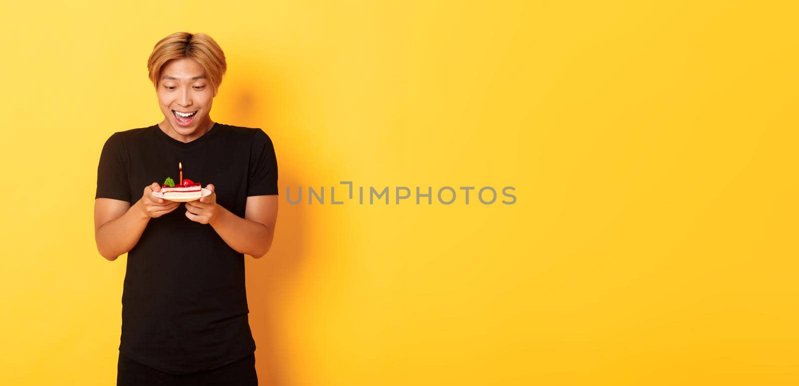 Excited happy attractive asian guy smiling as looking at birthday cake, making wish, celebrating b-day, standing yellow background by Benzoix