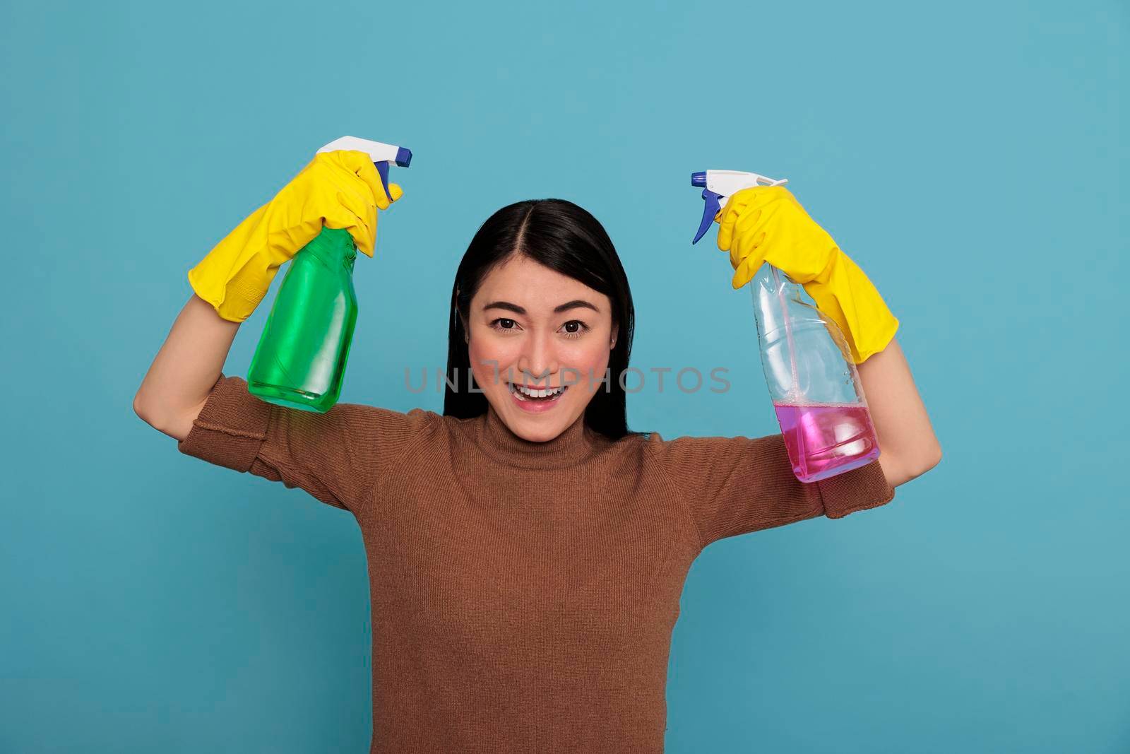 Excited glade and optimistic asian housekeeper at work raising arms holding two detergent sprays in the yellow gloves, Cleaning home concept, Cheerful pleased and positive housewife from chores