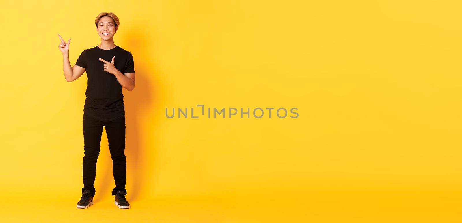 Full-length of smiling attractive korean guy pointing fingers left, showing logo, yellow background.