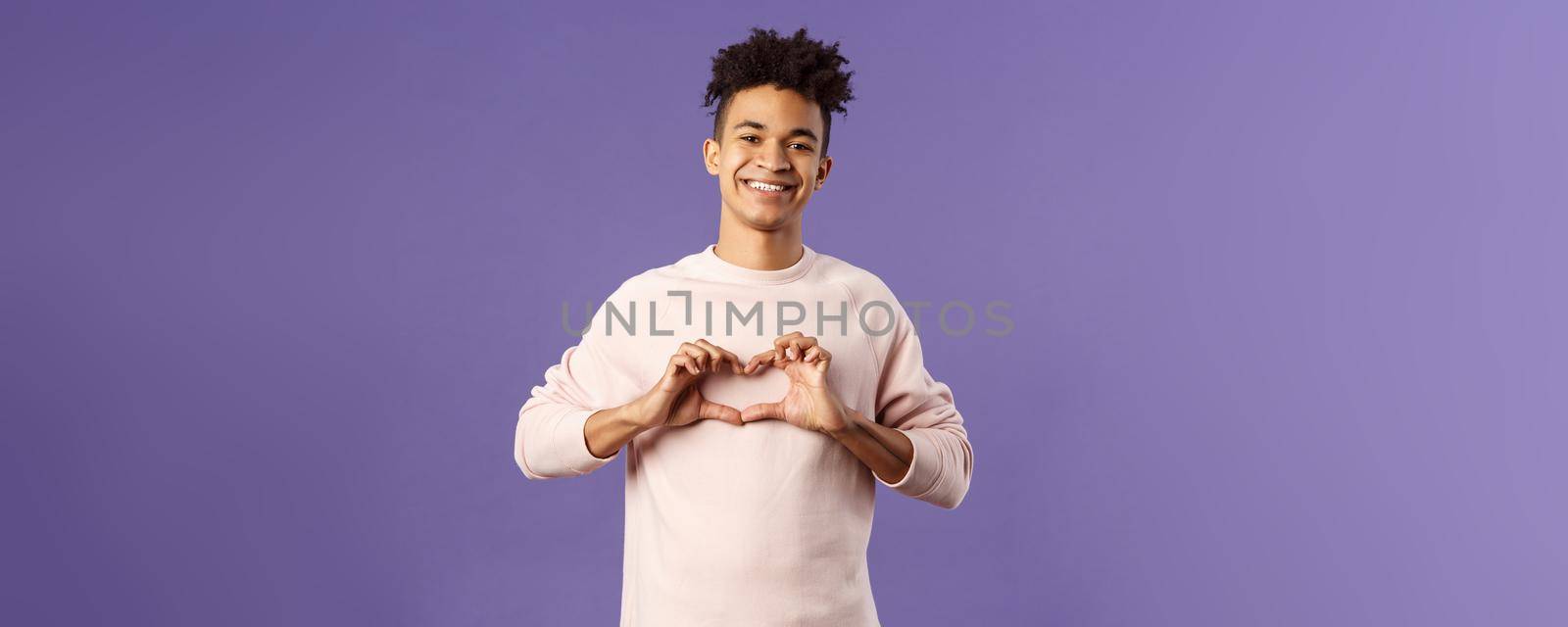 Portrait of young happy, romantic lovely boyfriend express his love and sympathy to dearest person, show heart sign and beaming smile, like this girl and trying to confess feelings, purple background.