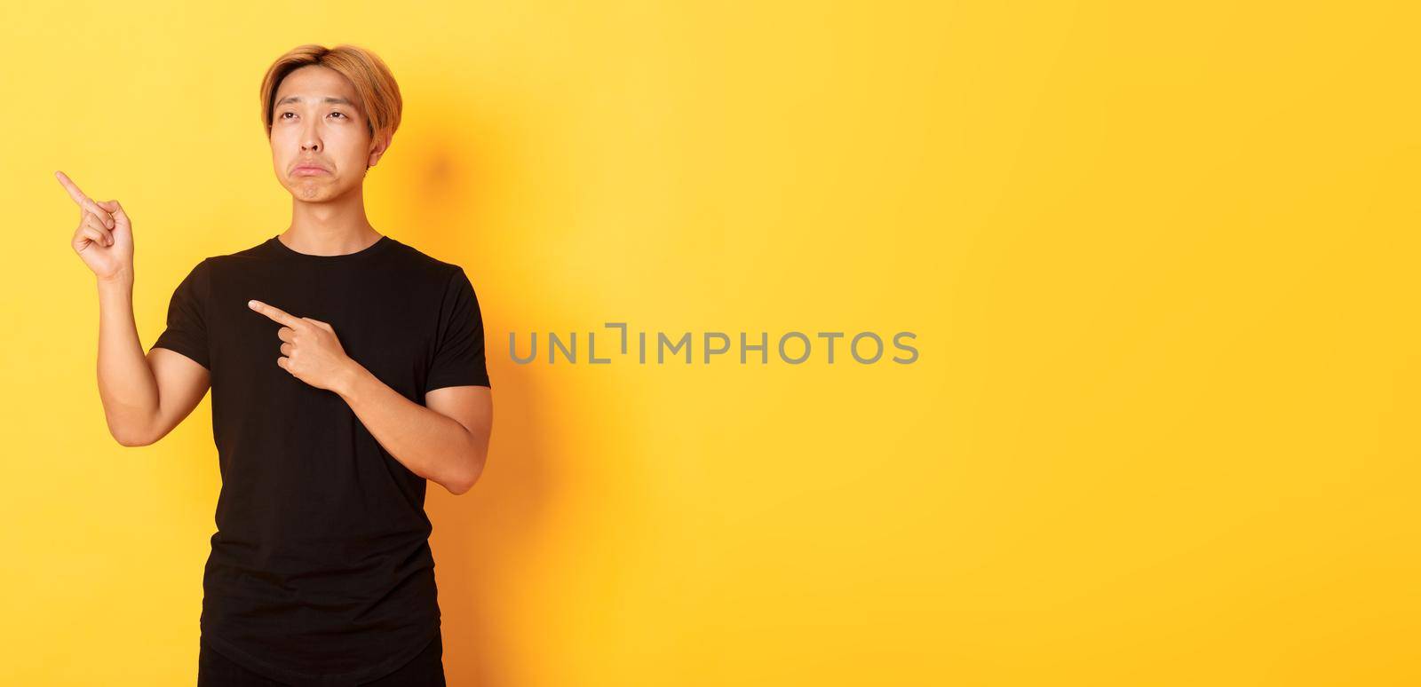 Portrait of pouting upset and funny asian guy pointing and looking upper left corner with regret, standing yellow background.