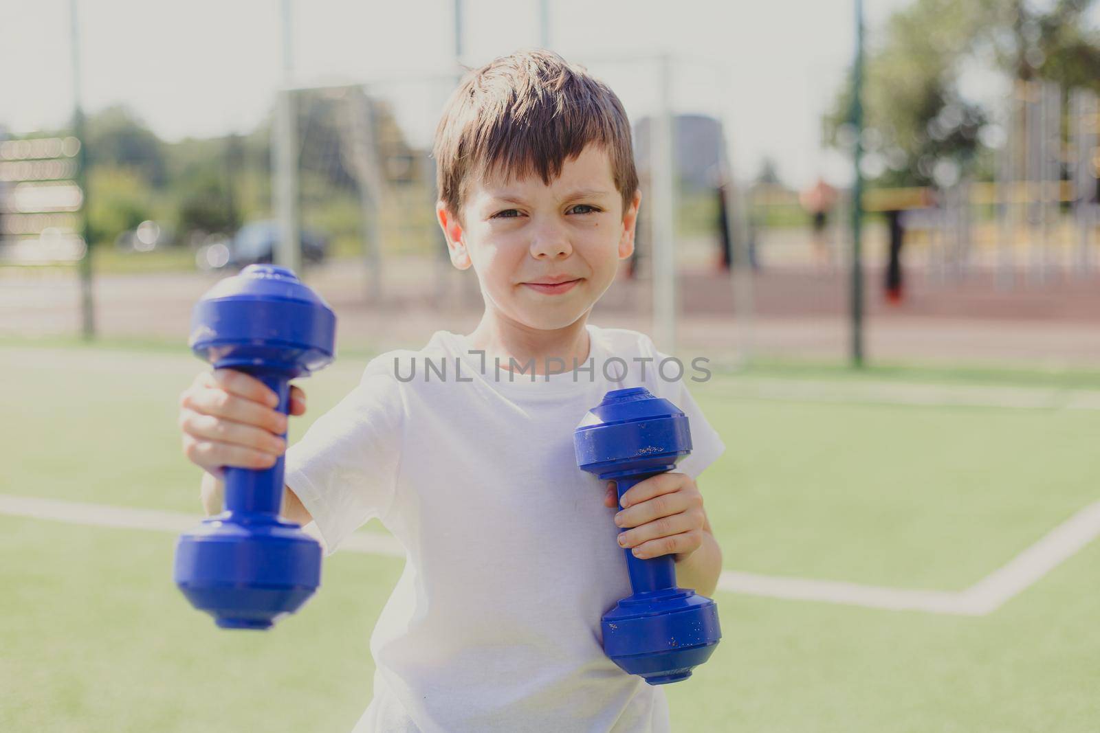 A child with dumbbells on the playground. A sporty kid. Illustrating an article about sports. Children's exercises. Education of champions. Light weightlifting. by alenka2194